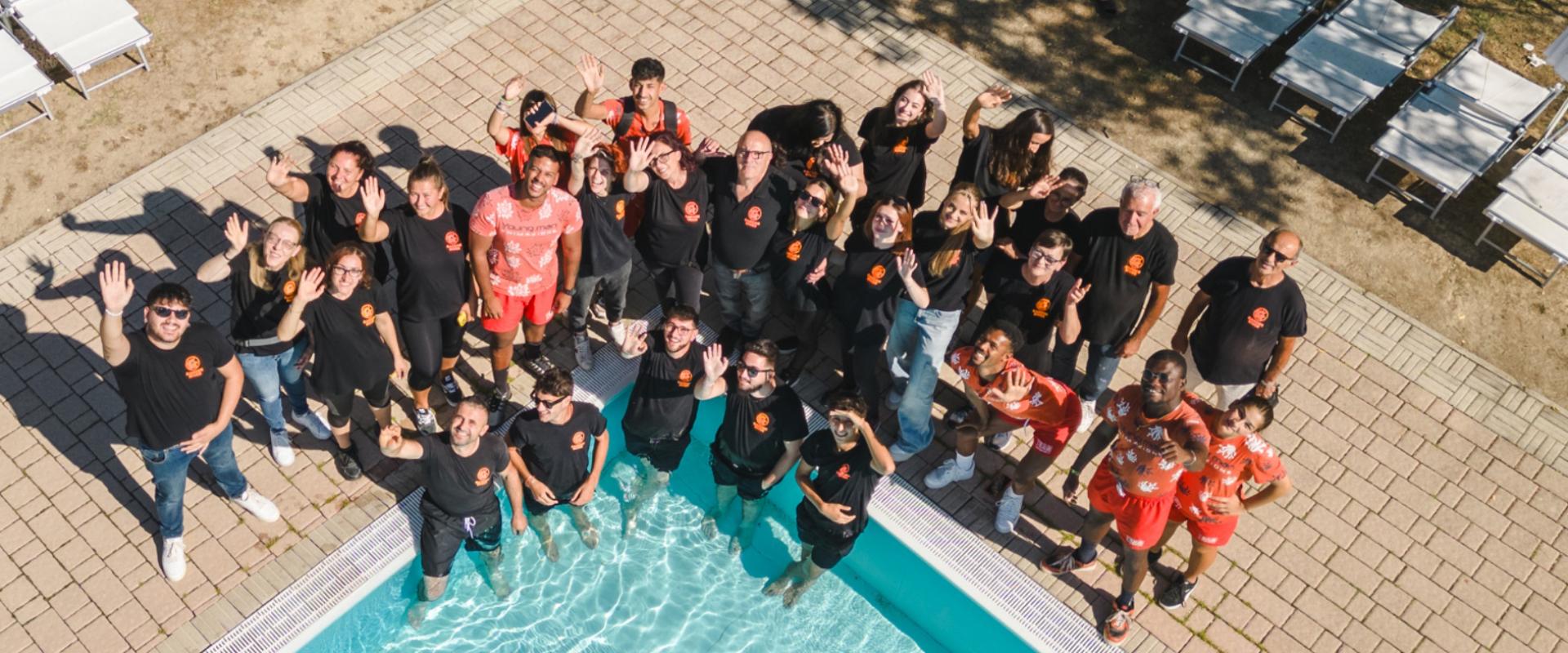 Gruppo di persone sorridenti vicino a una piscina, alcuni con magliette nere e altri rosse.
